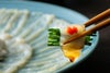 豊後とらふぐの刺身（てっさ）で刻みネギを巻いて、特製ポン酢につけているアップ画像。身の締まったふぐ刺しの食感と風味が伝わる、大分からのお取り寄せふぐ。