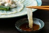 豊後とらふぐの刺身を、付属の特製ポン酢につけているアップ画像。ふぐ刺しの美しい身の締まりと、薬味の風味が伝わる、大分からのお取り寄せ品。