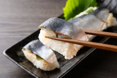 大分県産の「関さば棒寿司」（冷凍）を切って、一切れを箸で持ち上げたアップ画像。身の締まった関さばとシャリのバランスが伝わる、お取り寄せ寿司。