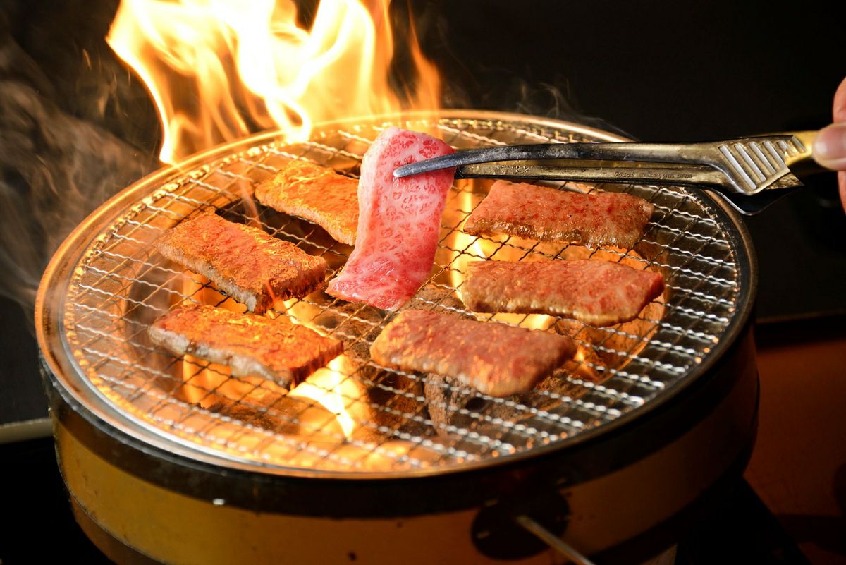 大分県産「おおいた和牛」のカルビまたはロースの切り身を、いろり（囲炉裏）の上で焼いている調理中の画像。高級和牛の脂が溶け出し、食欲をそそる焼肉風景。