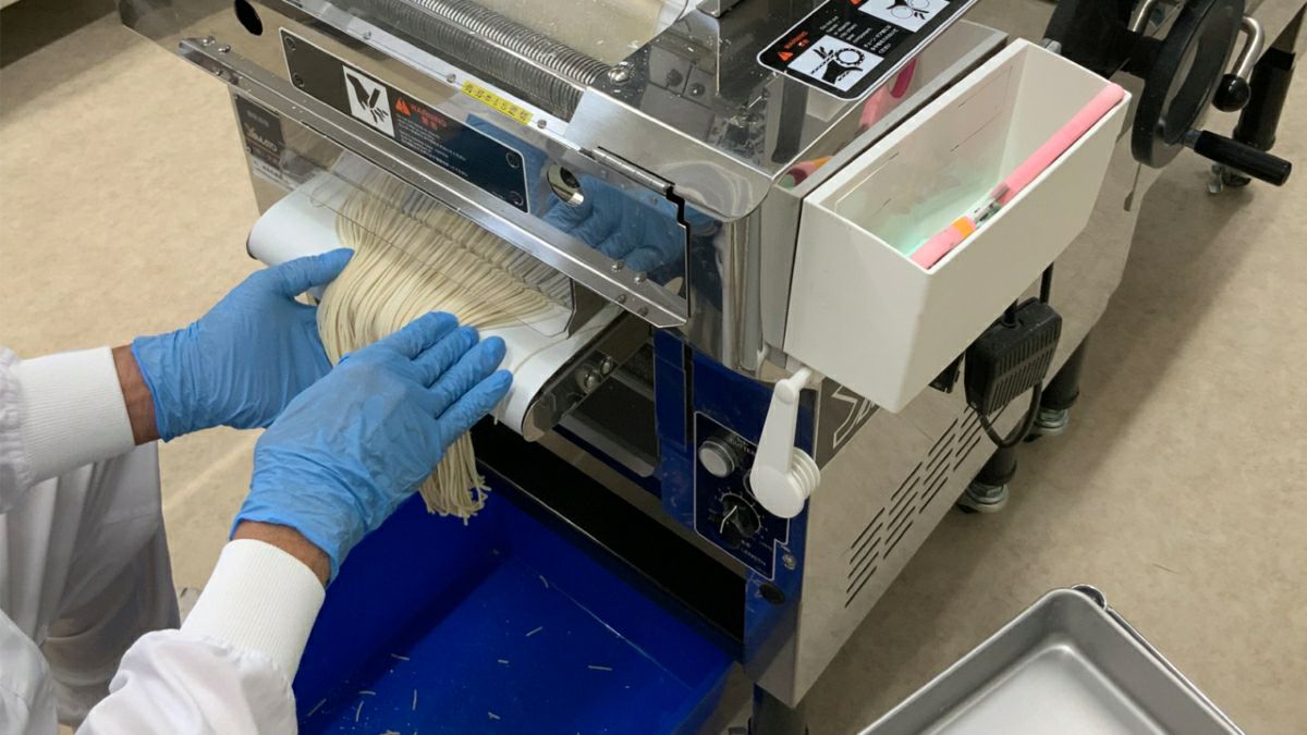 「別府温泉冷麺」の特製麺が製麺機から出てくるのを、職人の手が受け止めている画像。コシの強さを生み出す、こだわりの麺作り工程。