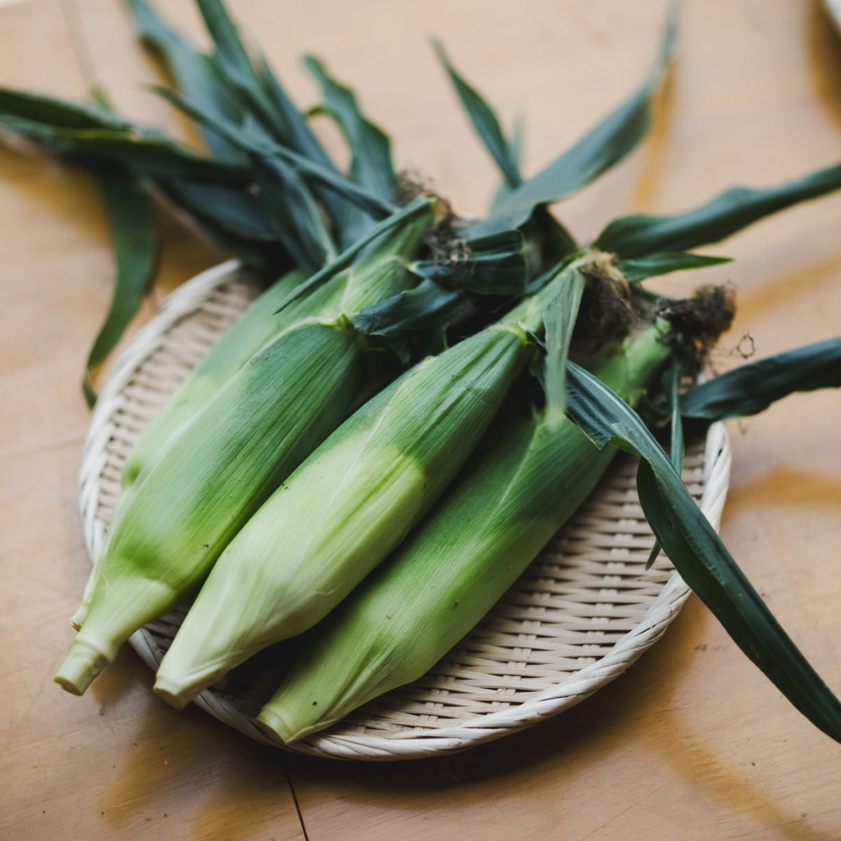 竹ざるに並べられた収穫したての皮付きとうもろこし。産地直送ならではの鮮度感溢れるイメージ