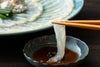 透き通るとらふぐの刺身を1枚箸で持ち上げ、特製ポン酢につける瞬間。豊後水道の荒波で育ったふぐならではの、引き締まった身の透明感を伝えるシズル写真。