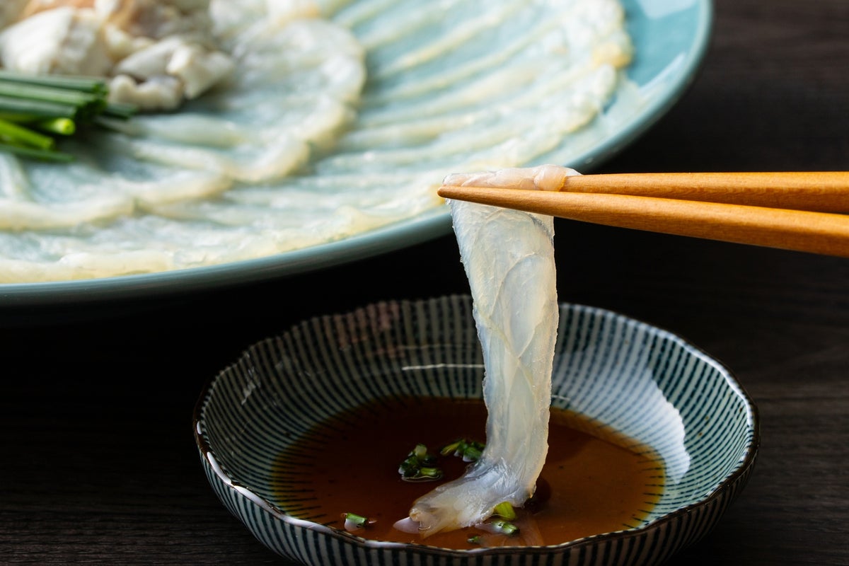 透き通るとらふぐの刺身を1枚箸で持ち上げ、特製ポン酢につける瞬間。豊後水道の荒波で育ったふぐならではの、引き締まった身の透明感を伝えるシズル写真。